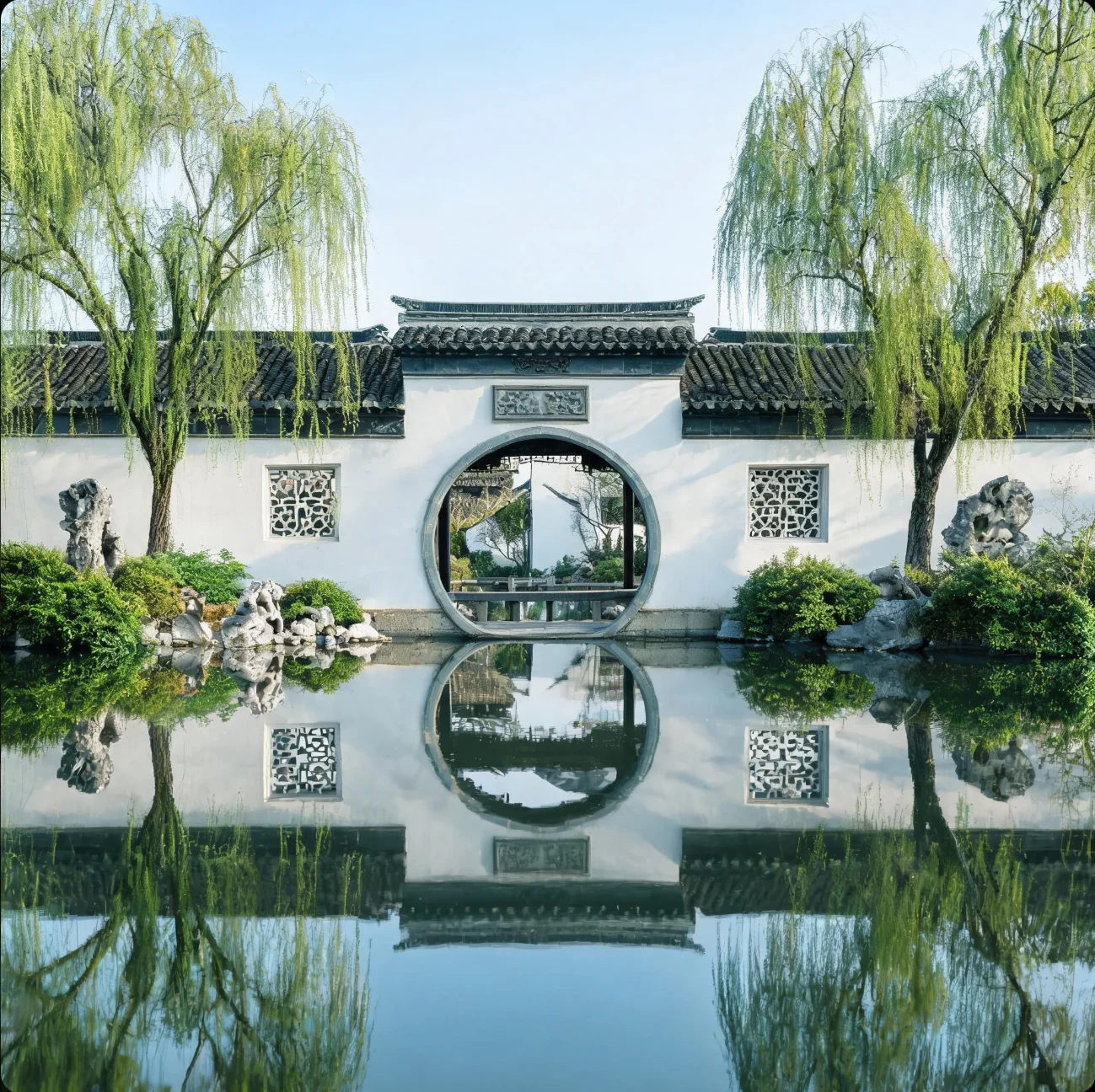白墙圆门与两侧垂柳临水而立,墙面、树木和门洞的倒影映在池中。